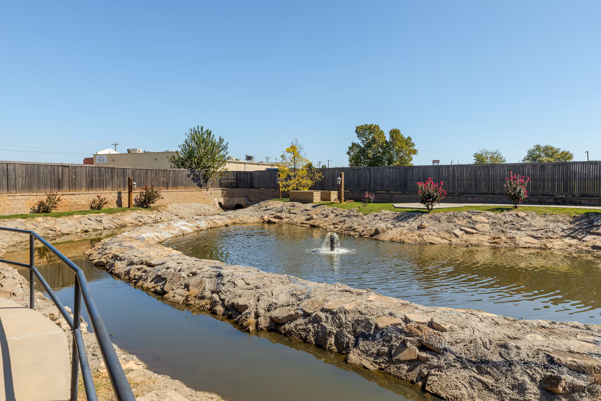 Outdoor area with a small pond featuring a central water fountain, surrounded by rocky edges and a concrete walkway with a metal railing. There are small trees and flowering bushes planted along the perimeter, with a wooden fence and some buildings visible in the background under a clear blue sky.