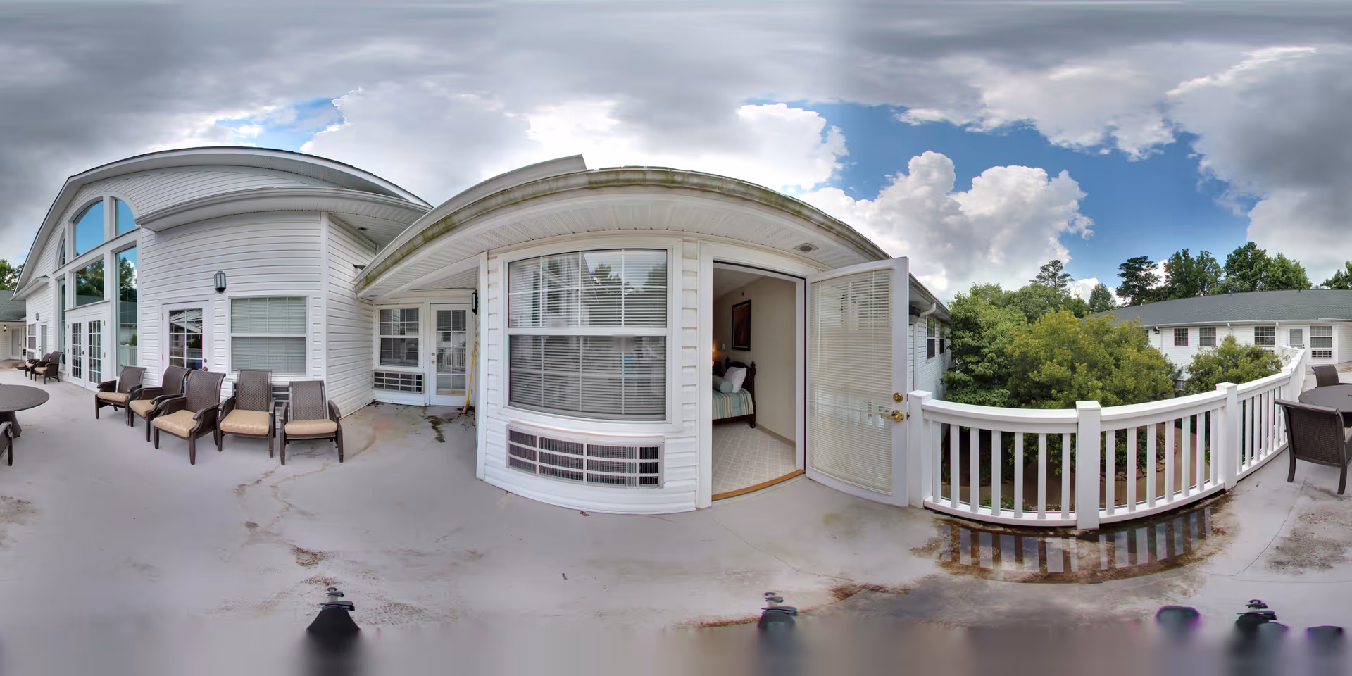 Outdoor patio area at Oaks at Tucker with several chairs arranged along the building's white exterior wall. A door is open leading into a bedroom with a bed visible inside. The patio has a white railing overlooking green trees and another building in the background under a partly cloudy sky.