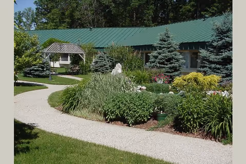 Landscaped courtyard with a winding gravel path, shrubs and small evergreen trees in front of a single-story building with a green metal roof.