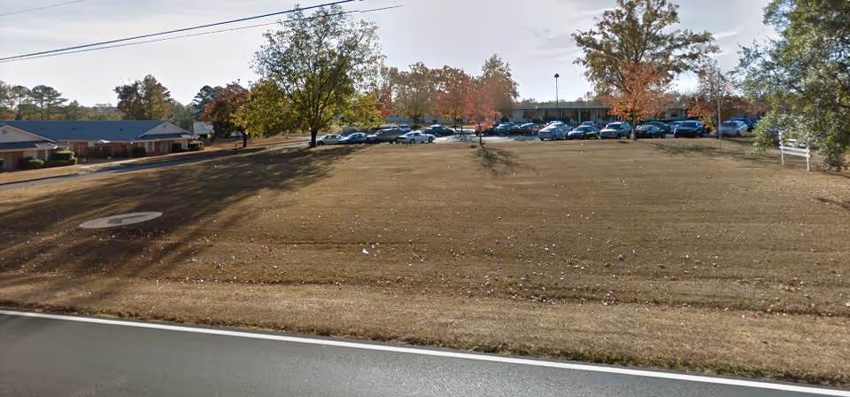 Wide view of a grassy lawn with scattered trees showing autumn foliage, a parking lot filled with cars, and a single-story building in the background under a partly cloudy sky.