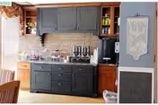 Interior view of a kitchen area with a black cabinet and countertop holding various kitchen appliances and items. The wall behind has a light brick backsplash, and there are wooden cabinets above and to the side. A decorative wall hanging is visible on the right side.
