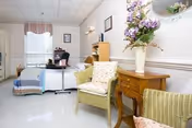 A bright and clean room with a single bed covered with a colorful quilt, a small desk with a chair, two wicker chairs with cushions, a wooden side table with a vase of flowers, and a window with curtains letting in natural light.