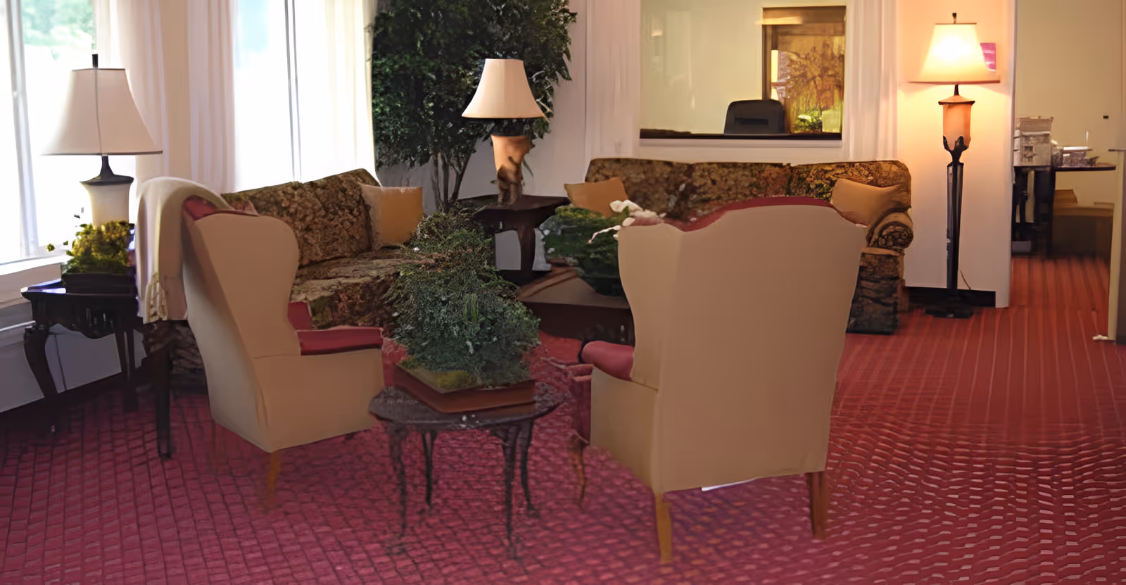 Well-lit common living room with upholstered armchairs and sofas, side tables, lamps, and potted plants on a patterned red carpet.
