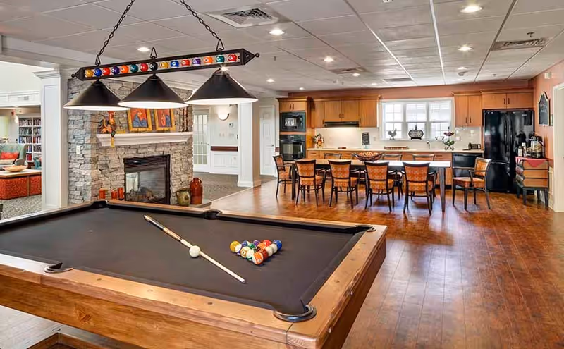 A spacious common area featuring a pool table with a cue stick and racked billiard balls in the foreground, a stone fireplace with framed artwork above it, and a large dining table with chairs in front of a kitchen area with wooden cabinets and black appliances.