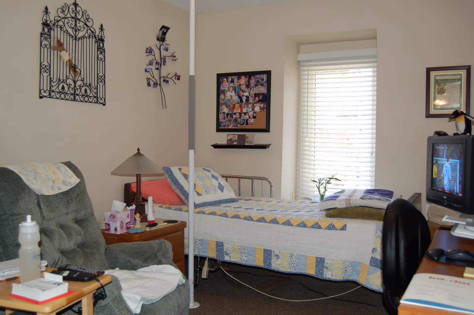 A cozy assisted living bedroom with a single bed covered in a patchwork quilt, a green recliner chair with a white cloth draped over the arm, a wooden nightstand with a lamp, tissues, and personal items, a window with white blinds letting in natural light, a TV on a desk showing a program, and decorative wall art including a metal wall hanging and a photo collage.
