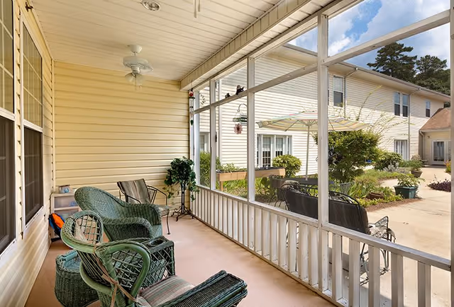 Screened-in porch area with green wicker chairs and a small table, overlooking a courtyard with plants and a building with white siding and multiple windows.