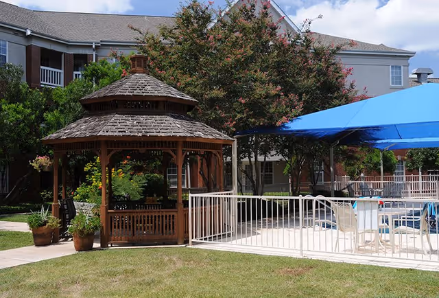 Outdoor area of a senior living facility featuring a wooden gazebo with plants around it, a fenced swimming pool with a blue canopy, white chairs and tables, and a multi-story building in the background surrounded by trees and greenery.