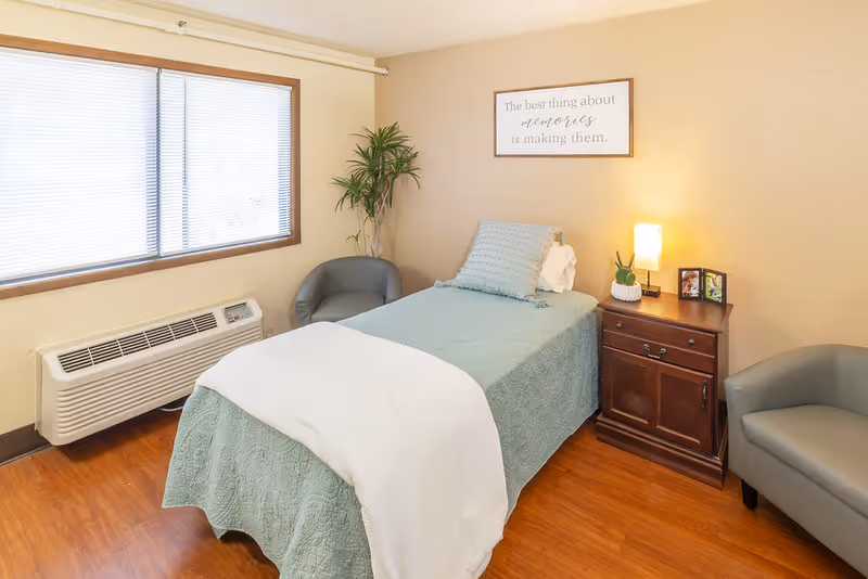 A cozy bedroom in an assisted living facility featuring a single bed with a light blue quilt and white blanket, two gray armchairs, a wooden nightstand with a small lamp, plant, and framed photos, a large window with blinds, and a framed quote on the wall that reads 'The best thing about memories is making them.'