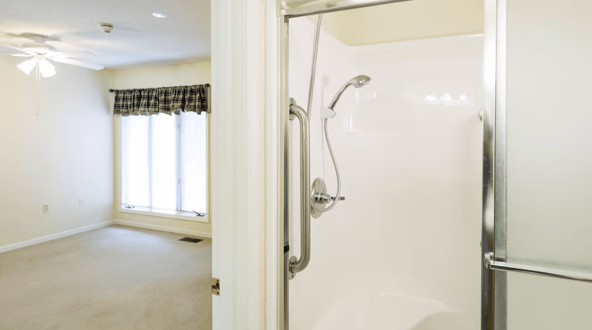 View of a bright, empty bedroom with beige carpet, a ceiling fan with lights, and a window with blinds and a plaid valance. Adjacent to the bedroom is a bathroom with a white shower stall featuring a handheld showerhead and a metal grab bar.