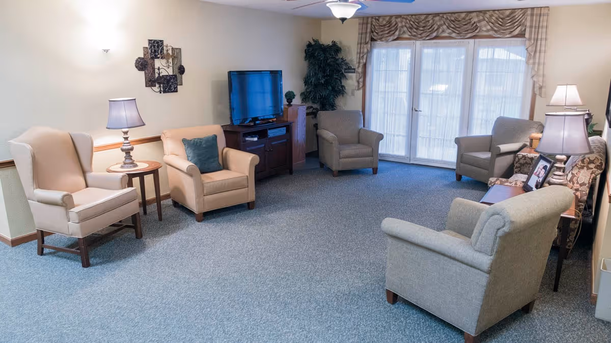 A cozy living room with five armchairs arranged around a central area. There is a wooden TV stand with a flat-screen television, a small round side table with a lamp, decorative wall art, and a large window with sheer curtains allowing natural light to enter the room.