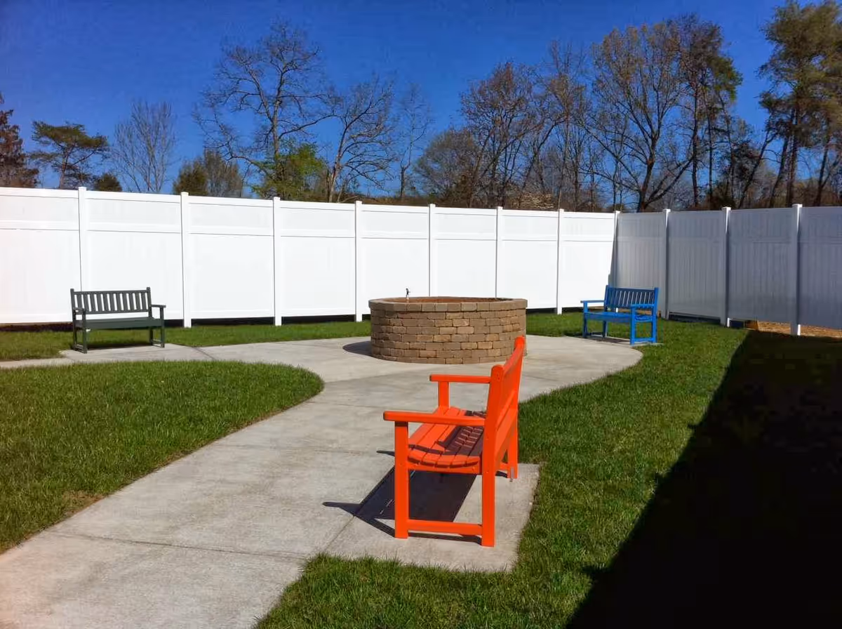 Outdoor courtyard area with a circular brick fire pit in the center, surrounded by a concrete walkway and three benches in different colors (red, blue, and green). The area is enclosed by a white privacy fence and trees are visible in the background under a clear blue sky.