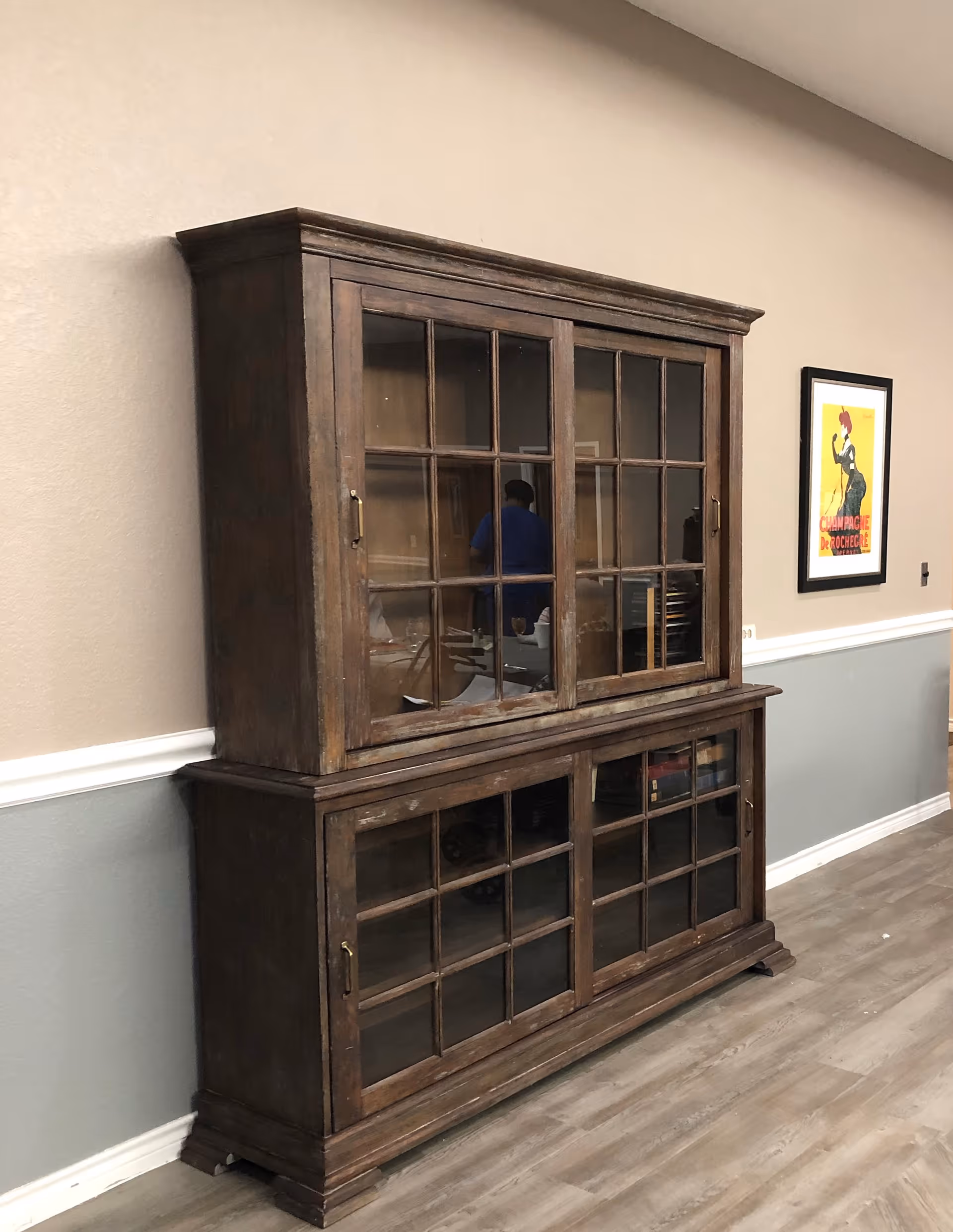 Large wooden glass-front cabinet against a beige wall in an interior hallway with framed art and wood-look flooring.