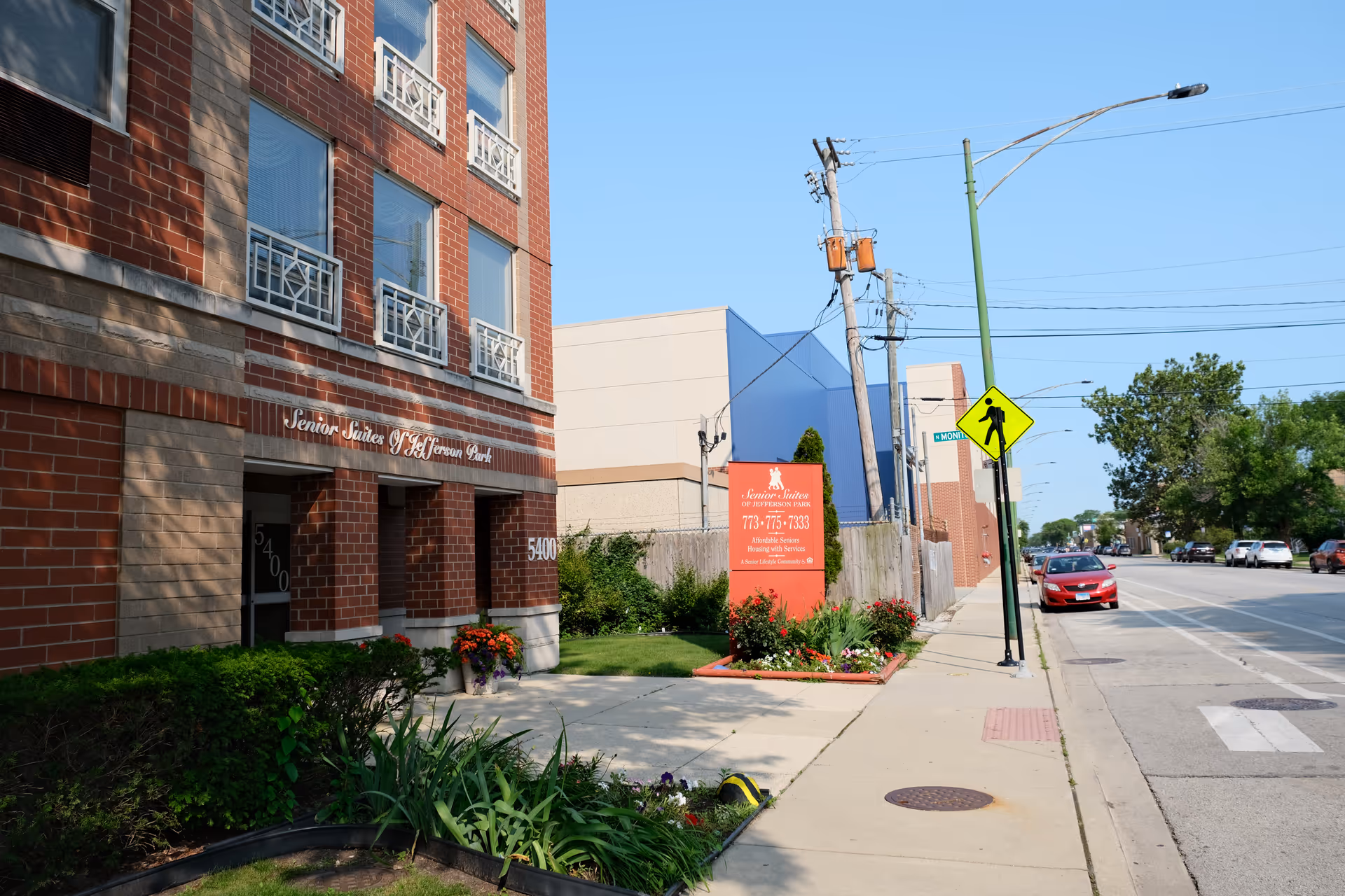 Exterior view of the Senior Suites of Jefferson Park building showing the brick facade, windows with decorative railings, and the entrance area with plants and flowers. A red sign with facility information is visible near the sidewalk along a street with parked cars and a pedestrian crossing sign.