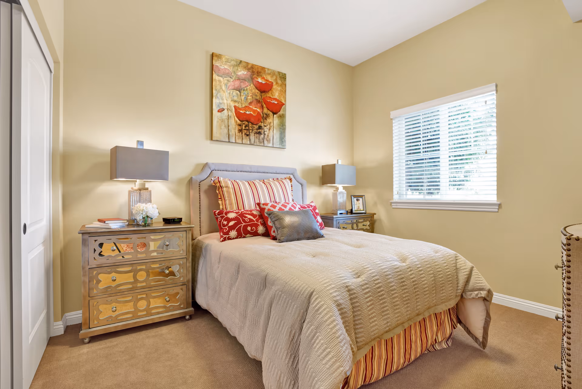 A cozy bedroom with a beige bedspread and multiple decorative pillows on a bed with a padded headboard. There are two mirrored nightstands on either side of the bed, each with a lamp and decorative items. A window with white blinds is on the right wall, and a painting of red flowers hangs above the bed. The walls are painted light beige, and the floor is carpeted.