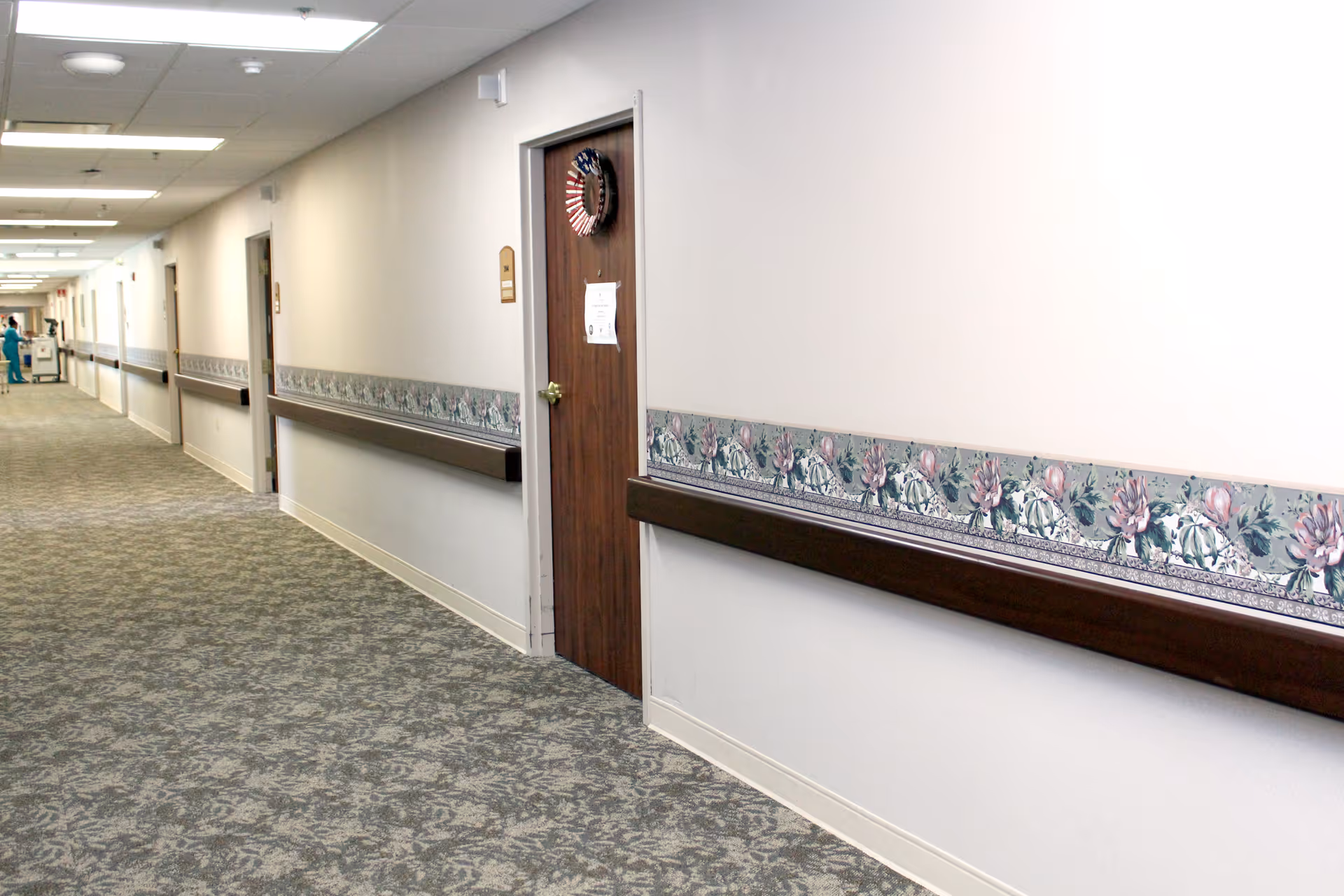 Long carpeted interior hallway of an assisted living facility with handrails, doors and a floral wall border.