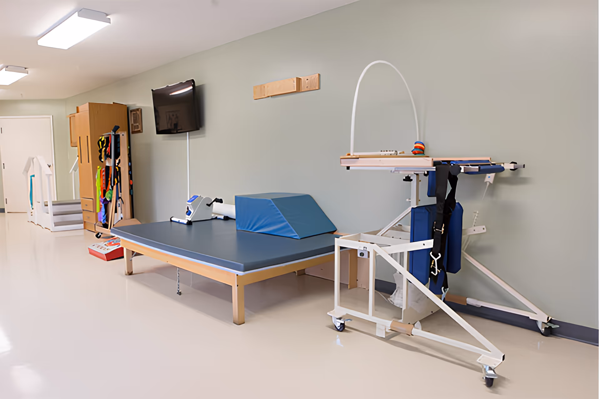A rehabilitation or physical therapy room with a padded therapy table, exercise equipment, a mounted TV, and various therapy aids against a light green wall.
