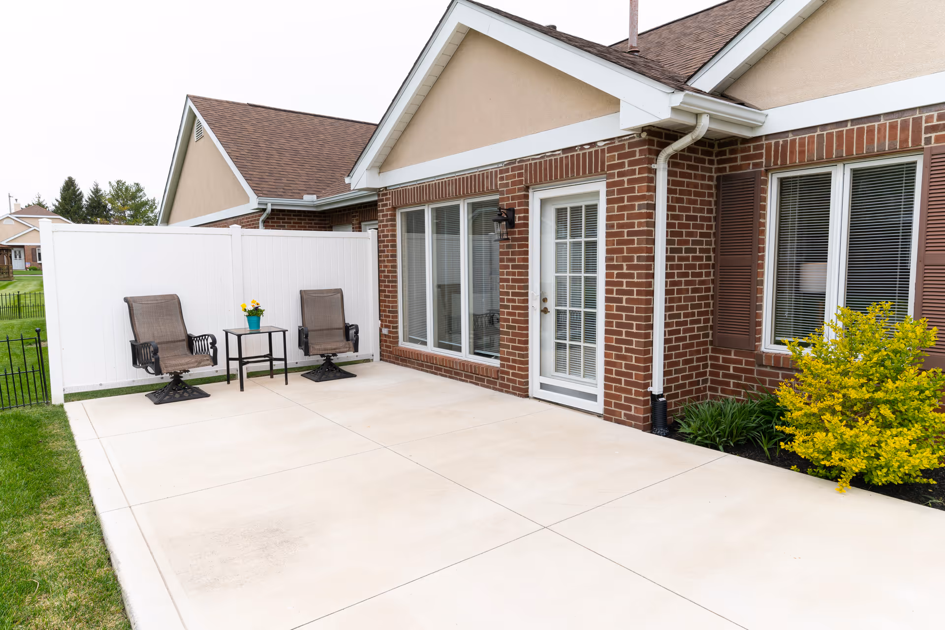 Outdoor patio with two chairs and a small table beside a brick building with glass doors and a white privacy fence.