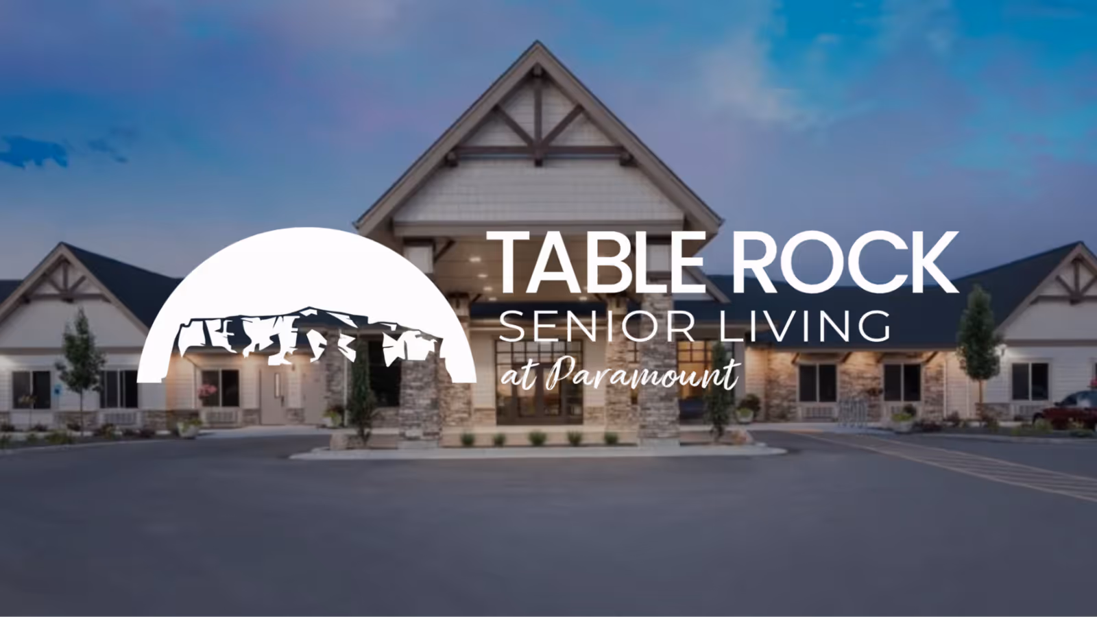 Exterior view of a senior living facility building with a peaked roof and stone pillars at the entrance, taken during dusk with a clear sky.