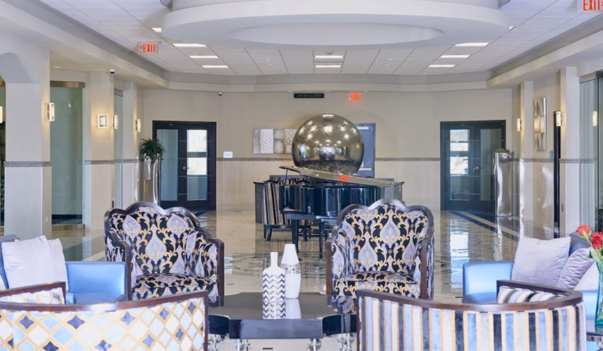 A spacious and elegant common area with patterned armchairs and sofas arranged around a black coffee table with decorative vases. In the background, there is a grand piano with a large reflective silver sphere on top. The room has a polished tile floor, light-colored walls, and multiple doors leading to other areas. Ceiling lights and wall sconces provide illumination.