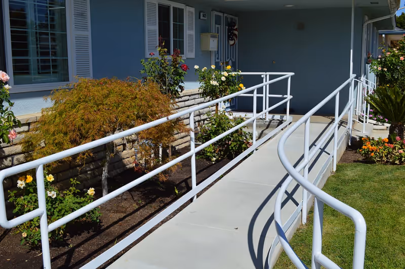 Concrete wheelchair ramp with white metal handrails leading to the front entrance of a single-story building bordered by flowers and shrubs.