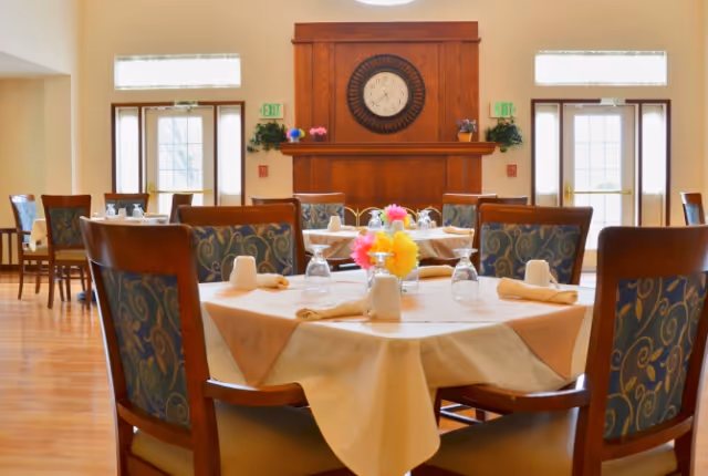 A bright dining room with round tables set with napkins and glasses, upholstered chairs, and a wooden mantel with a large clock.