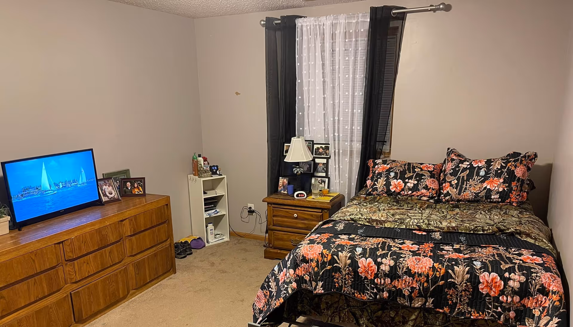 A bedroom with a bed covered in floral and camouflage patterned bedding, a wooden nightstand with a lamp, framed photos, and a digital clock. There is a window with sheer white curtains and black drapes behind the nightstand. On the left side of the room, there is a wooden dresser with a TV displaying a sailboat scene and several framed photos on top. The walls are plain and light-colored, and the floor is carpeted.