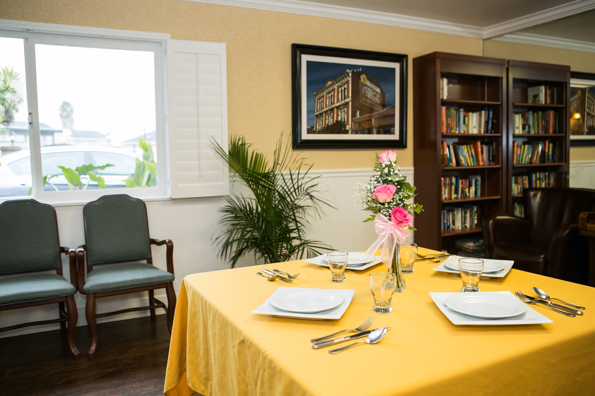 A dining table with a yellow tablecloth set for four and a pink floral centerpiece in a cozy common room with bookshelves and chairs.