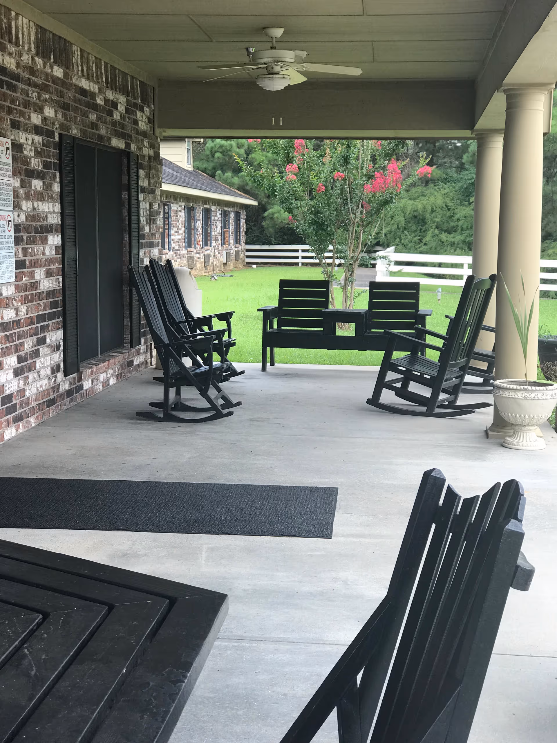 Covered outdoor patio area with black rocking chairs and benches arranged around a concrete floor. The patio is attached to a brick building with black shutters on the windows. There is a ceiling fan above and a green lawn with trees and a white fence visible in the background.