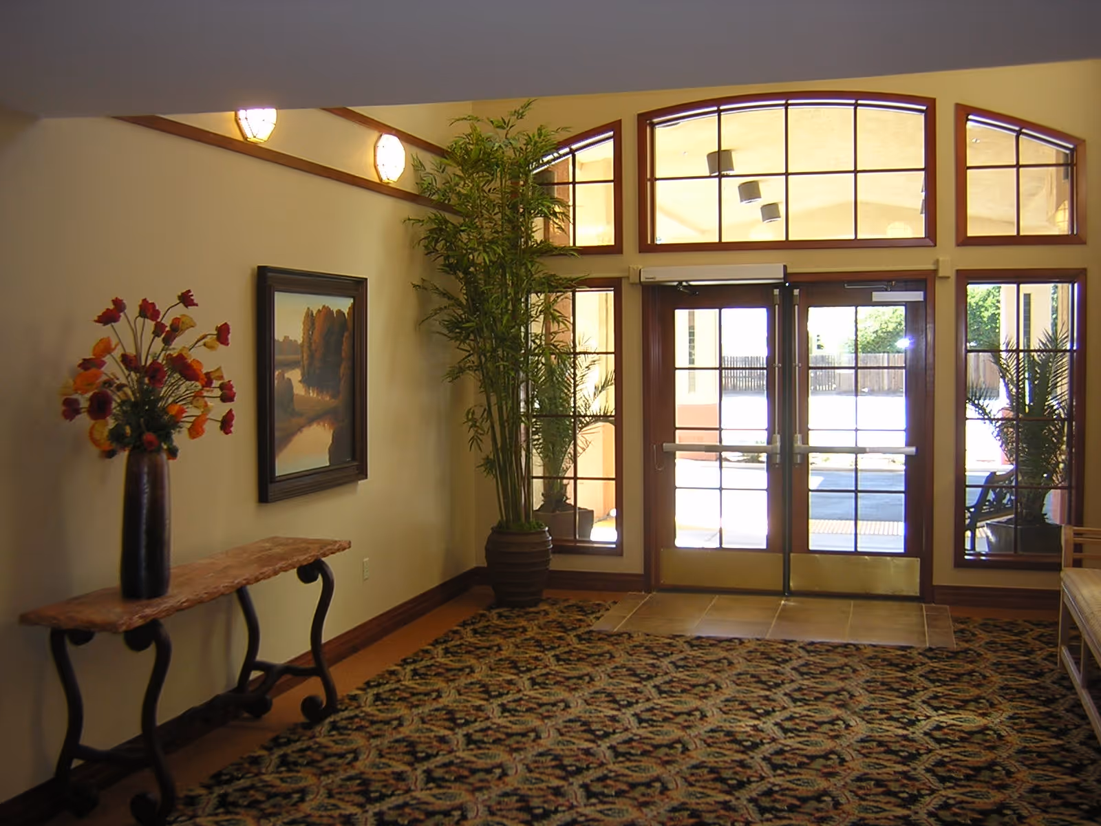 Entrance area of a retirement community with double glass doors and large windows above. The space features a patterned carpet, a wooden console table with a vase of flowers, a tall potted plant, and a framed landscape painting on the wall.