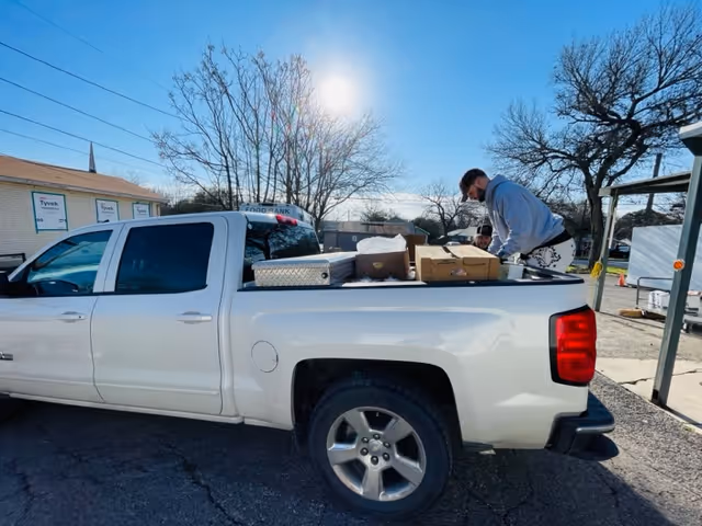 A white pickup truck parked outdoors with boxes in its open bed and a person leaning over them under a sunny sky.