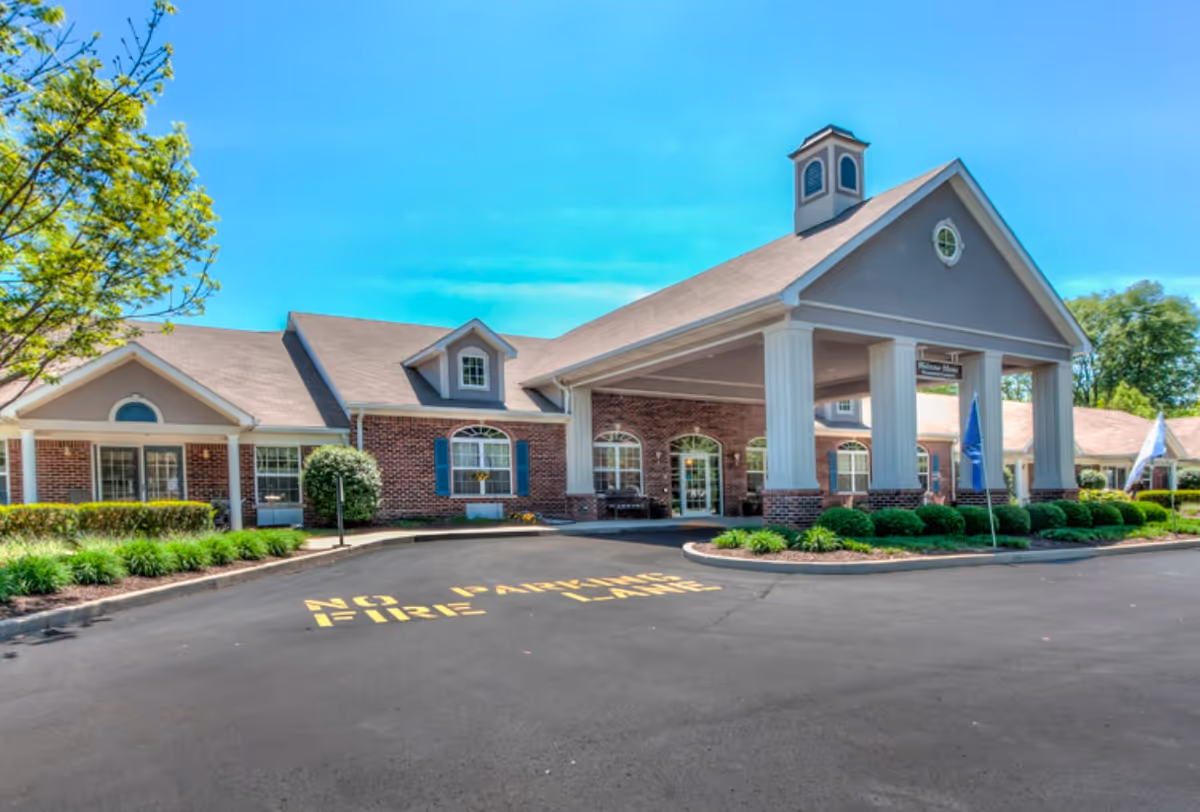 Exterior view of Five Star Residences of Noblesville showing a large brick building with a covered entrance supported by white columns. The building has multiple windows with blue shutters and a small cupola on the roof. There are landscaped bushes and trees around the building, and a paved driveway with 'NO PARKING FIRE LANE' painted in yellow on the asphalt.