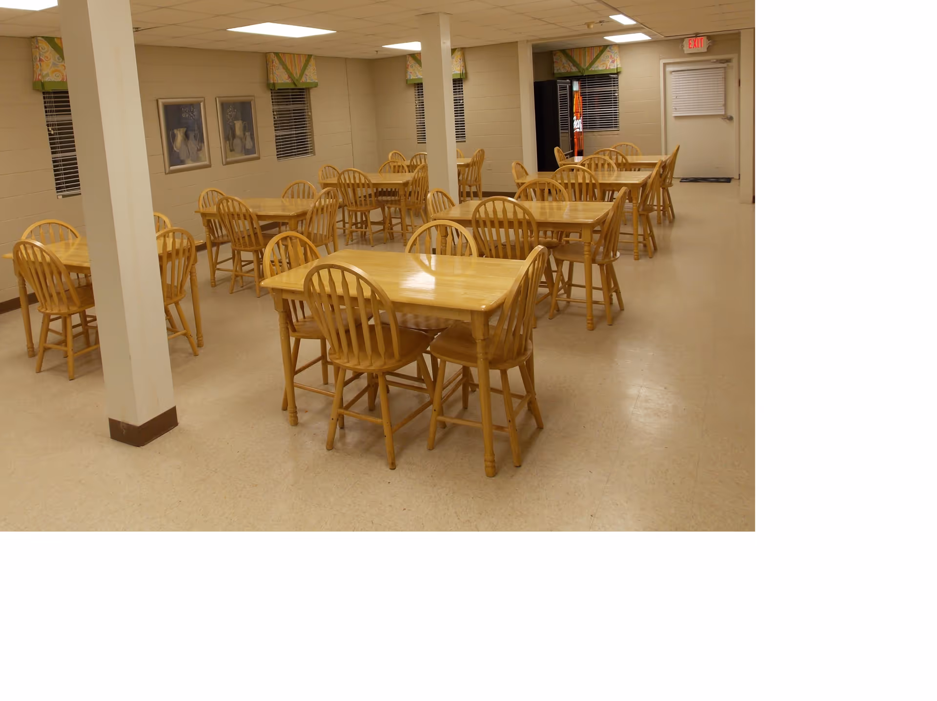 A dining room with multiple wooden tables and chairs arranged neatly. The room has beige walls with small windows covered by patterned valances. There are two framed pictures on one wall and a vending machine near an exit door at the back.