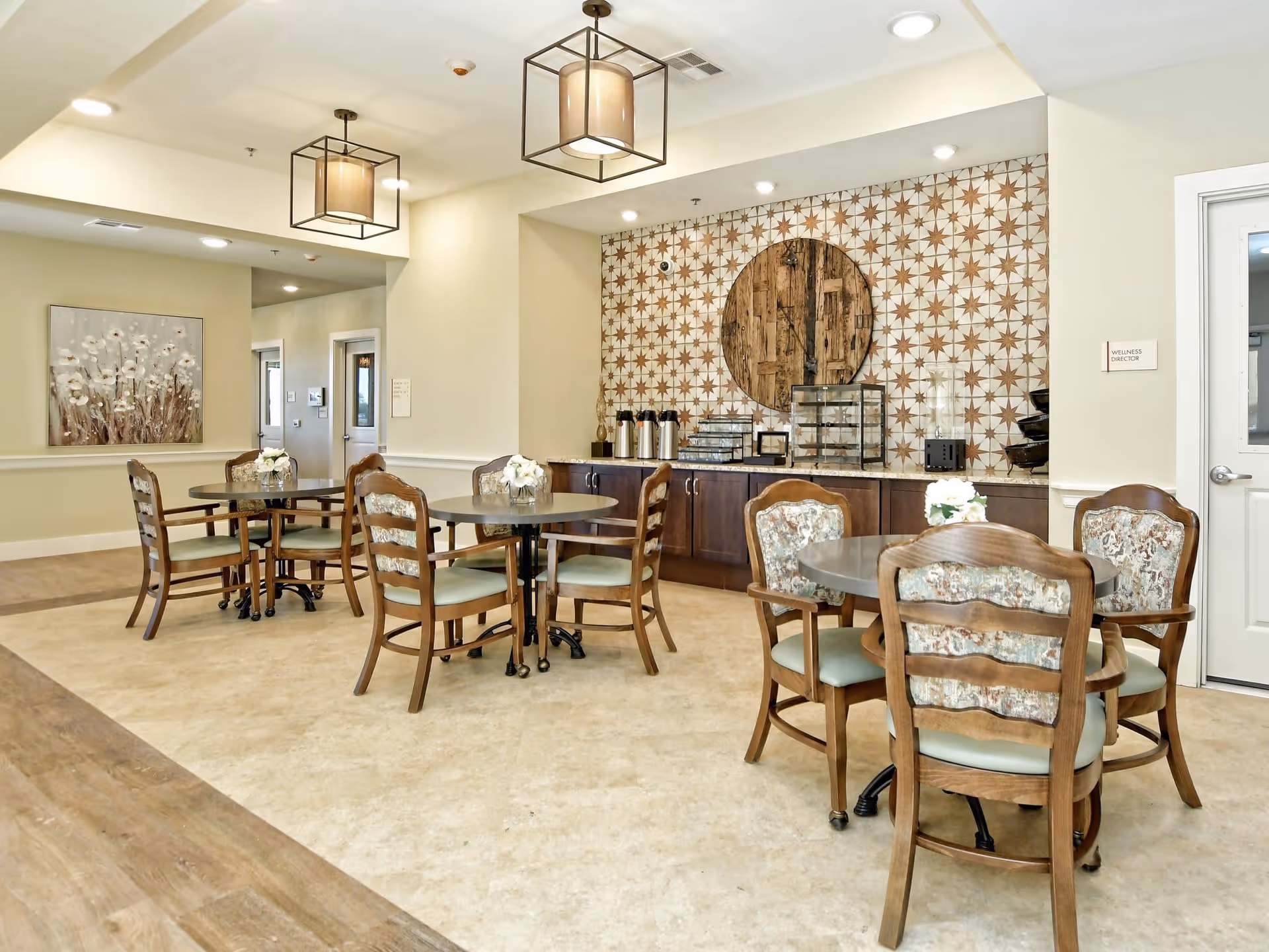 A bright and clean dining area with round tables and wooden chairs featuring floral upholstery. The back wall has a decorative tile pattern with a large circular wooden accent. There are coffee dispensers and display cases on a counter beneath the tiled wall. The room has modern ceiling lights and a painting of flowers on one wall.