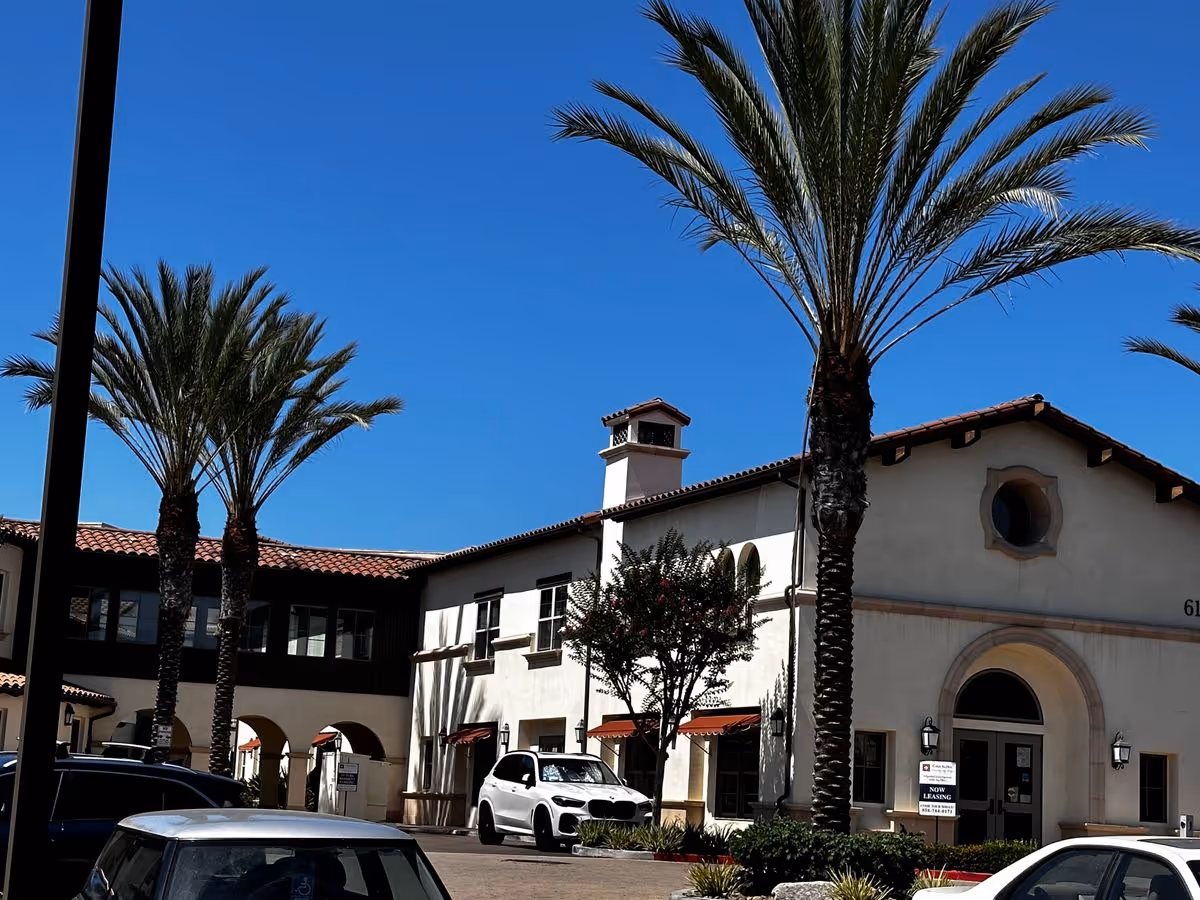 Mediterranean-style two-story building with palm trees and cars parked in front under a clear blue sky.