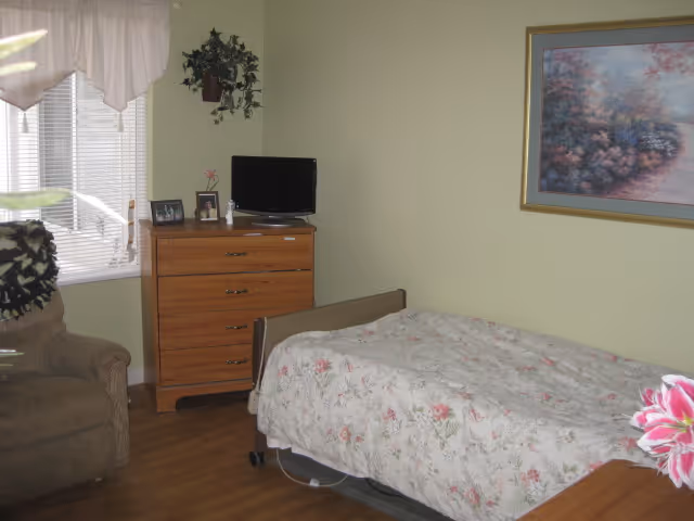 A cozy bedroom in an assisted living facility featuring a single bed with a floral bedspread, a wooden dresser with a small TV and framed photos on top, a comfortable armchair near a window with blinds and a valance, a potted plant hanging on the wall, and a framed landscape painting on the wall.