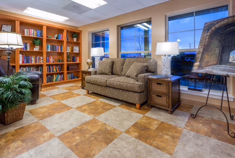 A cozy senior living facility common area with a patterned sofa, two side tables with lamps, a large bookshelf filled with books and decorative items, a leather armchair, a potted plant, and a large painting on an easel near three windows showing an outdoor view at dusk.