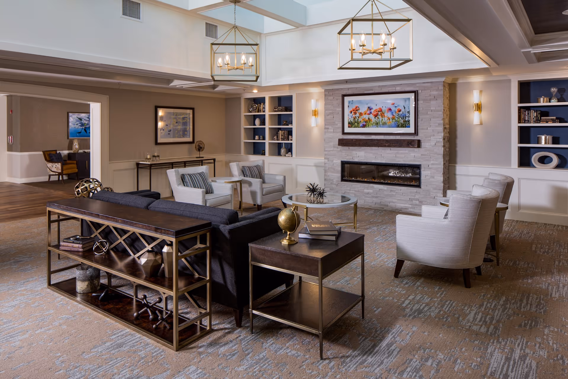 A spacious and well-lit living room area with a modern fireplace set in a stone wall. The room features a dark sofa, several light-colored armchairs, a glass coffee table, and side tables with decorative items including books and a globe. Built-in shelves flank the fireplace, and two large pendant lights hang from the ceiling. The room has a neutral color palette with blue accents and carpeted flooring.