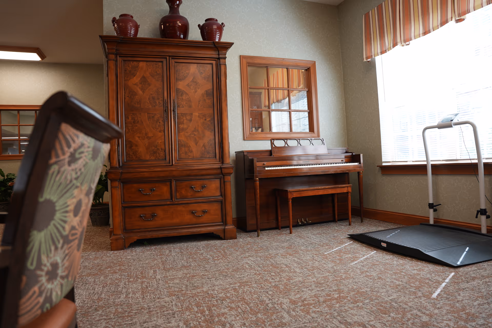 Cozy interior room with a wooden armoire, upright piano and bench beside a window and a treadmill mat, with an upholstered chair in the foreground.