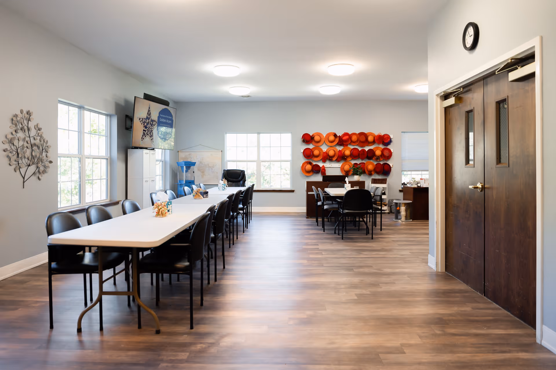 A spacious, well-lit room with long tables and black chairs arranged for group seating. The room has large windows letting in natural light, a wall-mounted TV, a water dispenser, and a decorative display of orange and red hats on the far wall. There are double wooden doors on the right side and a clock above them.