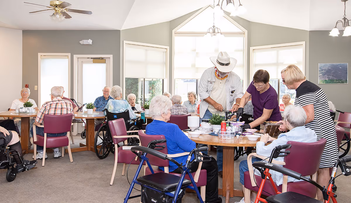 A group of elderly people sitting around tables in a bright room with large windows. Some are in wheelchairs and others are seated in chairs. Three caregivers are assisting and interacting with the residents. The room has ceiling fans and light fixtures, and the walls are painted gray with a few framed pictures.