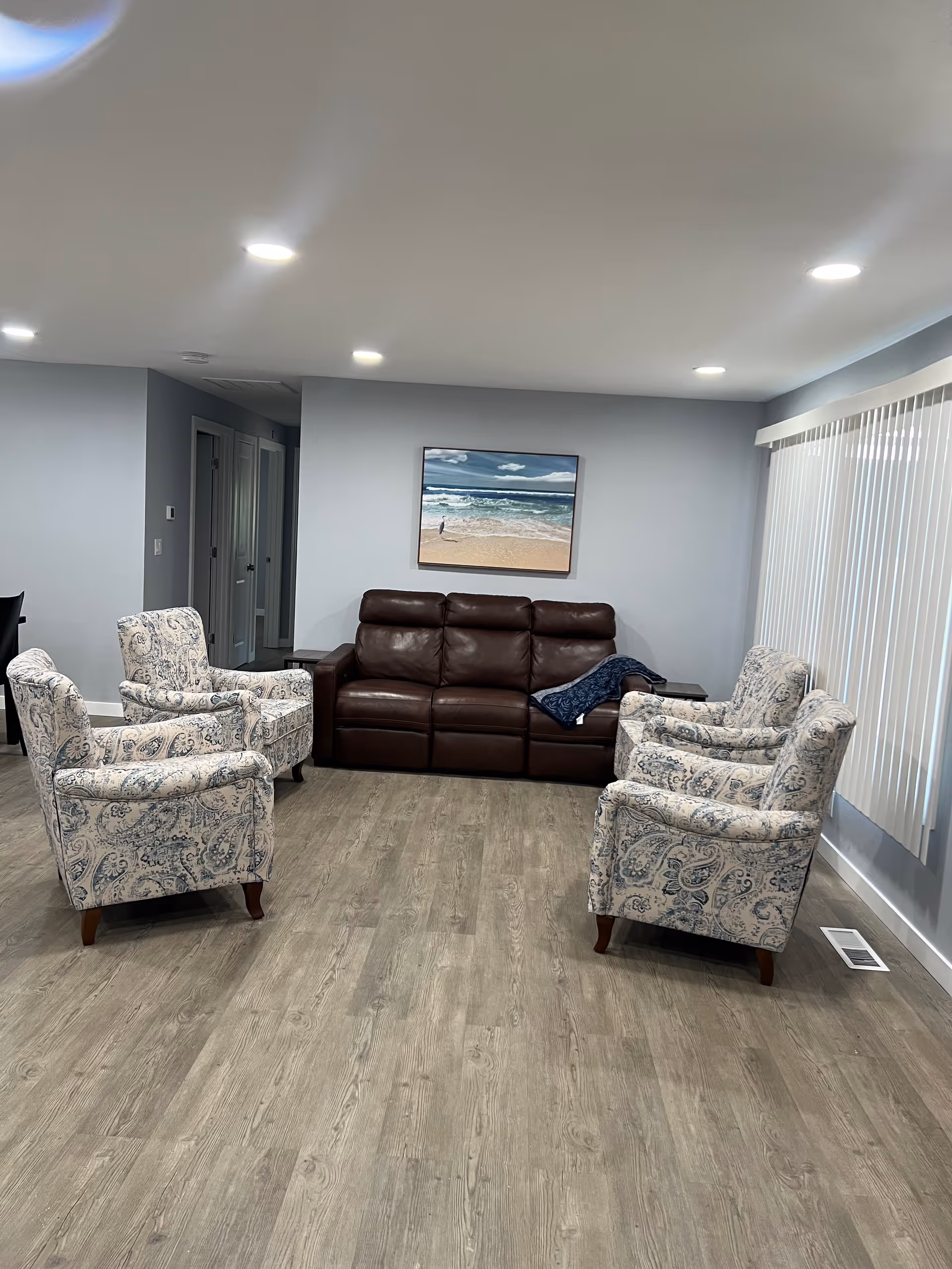 A living room with a brown leather sofa against a light gray wall, flanked by four patterned armchairs arranged in a semi-circle. A framed picture of a beach scene hangs above the sofa. The room has wood-look flooring and vertical blinds covering a large window on the right side. Recessed ceiling lights illuminate the space.
