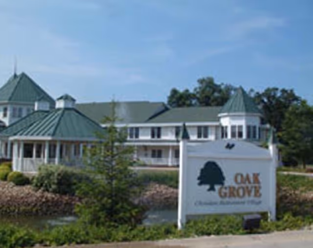 Front exterior of a multi-story retirement building with green roofs, landscaping, a small pond, and a white sign reading "OAK GROVE".