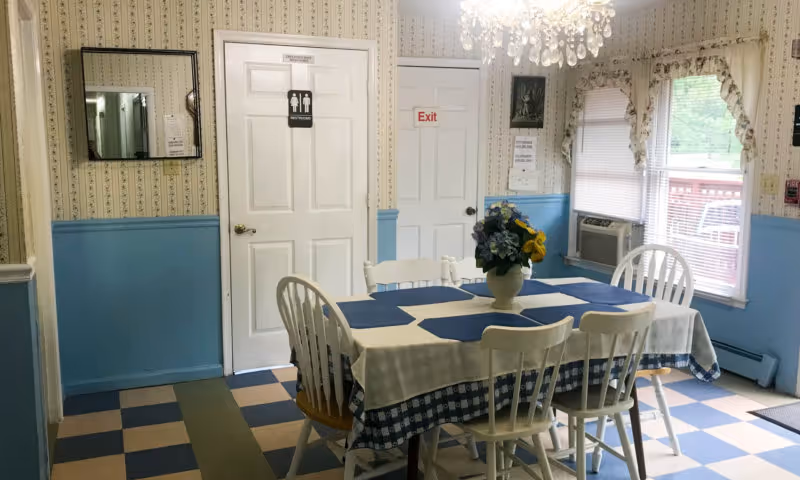 A small dining area with a rectangular table covered with a white and blue checkered tablecloth and blue placemats. Six white wooden chairs surround the table. A vase with flowers is placed in the center of the table. The room has blue and beige checkered flooring, wallpaper with a floral pattern on the upper half of the walls, and blue paint on the lower half. There is a window with blinds and floral curtains on the right side, an air conditioning unit below the window, and two white doors on the back wall, one labeled as a restroom and the other with an exit sign above it. A chandelier hangs from the ceiling.