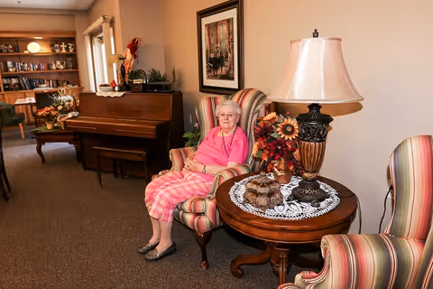 An elderly woman wearing a pink top and plaid pants sits in a striped armchair in a cozy living room area. Next to her is a round wooden table with a decorative lamp, a floral arrangement, and a doily. In the background, there is a piano, bookshelves, and another armchair.