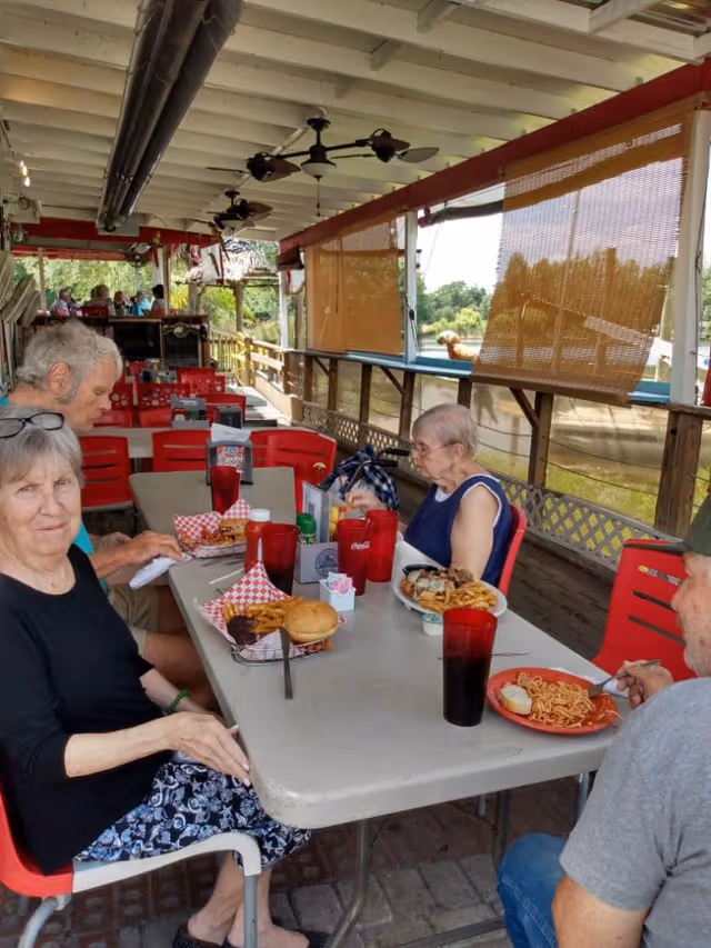 Four elderly people sitting around a table on a covered outdoor patio, eating meals including burgers, fries, and spaghetti. The patio has red chairs, ceiling fans, and roll-up bamboo shades with a view of greenery and water outside.