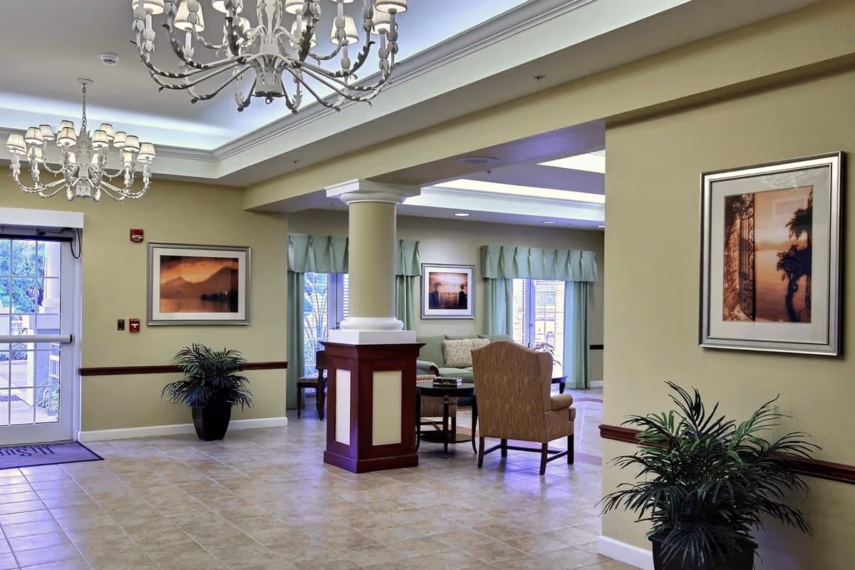 A spacious and well-lit interior common area of an assisted living community featuring tiled floors, beige walls with framed artwork, two chandeliers hanging from the ceiling, potted plants, and a seating area with chairs and a coffee table near large windows with green curtains.