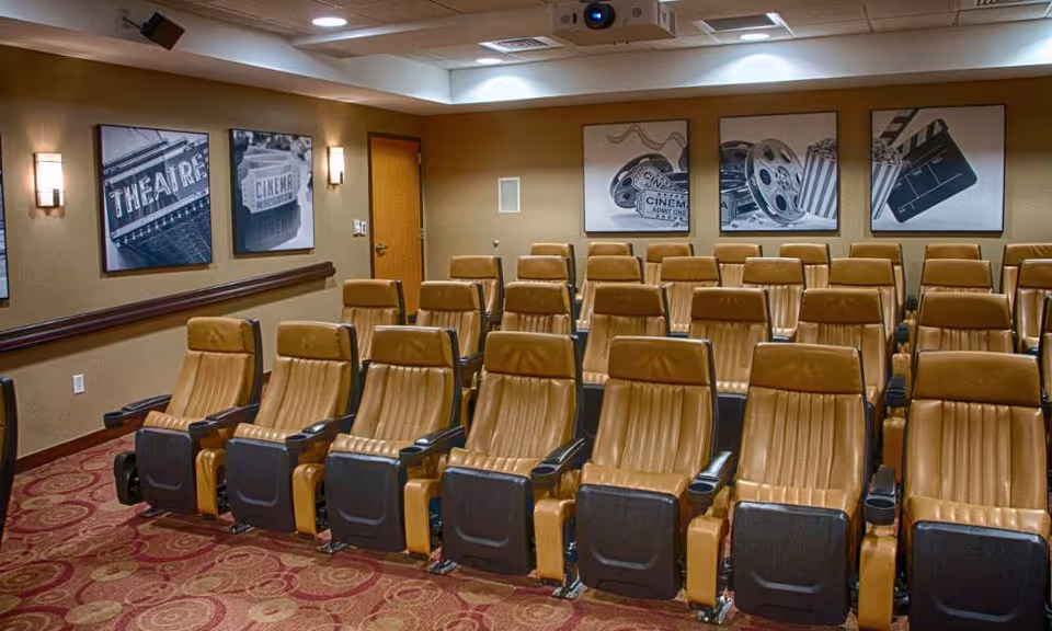 A small theater room with multiple rows of tan leather reclining seats arranged in front of a projector. The walls are decorated with black and white framed pictures related to cinema, including images of a theater marquee, film reels, popcorn, and a clapperboard. The room has warm lighting and a patterned carpet.