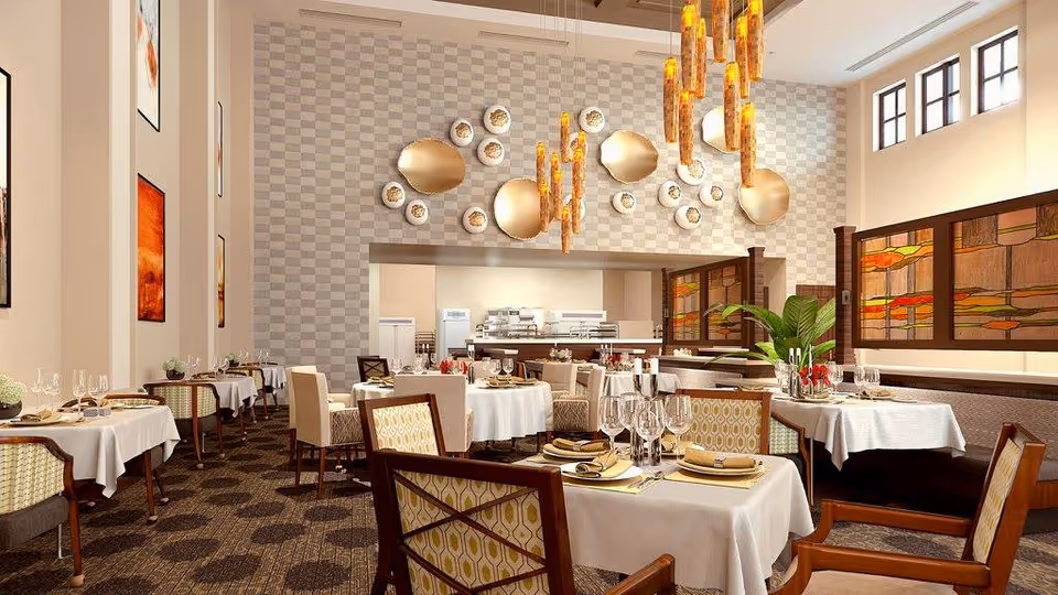 Elegant dining room with multiple tables covered in white tablecloths, set with plates, napkins, and glassware. The room features patterned carpet, decorative wall art, large windows with stained glass panels, and modern hanging light fixtures.