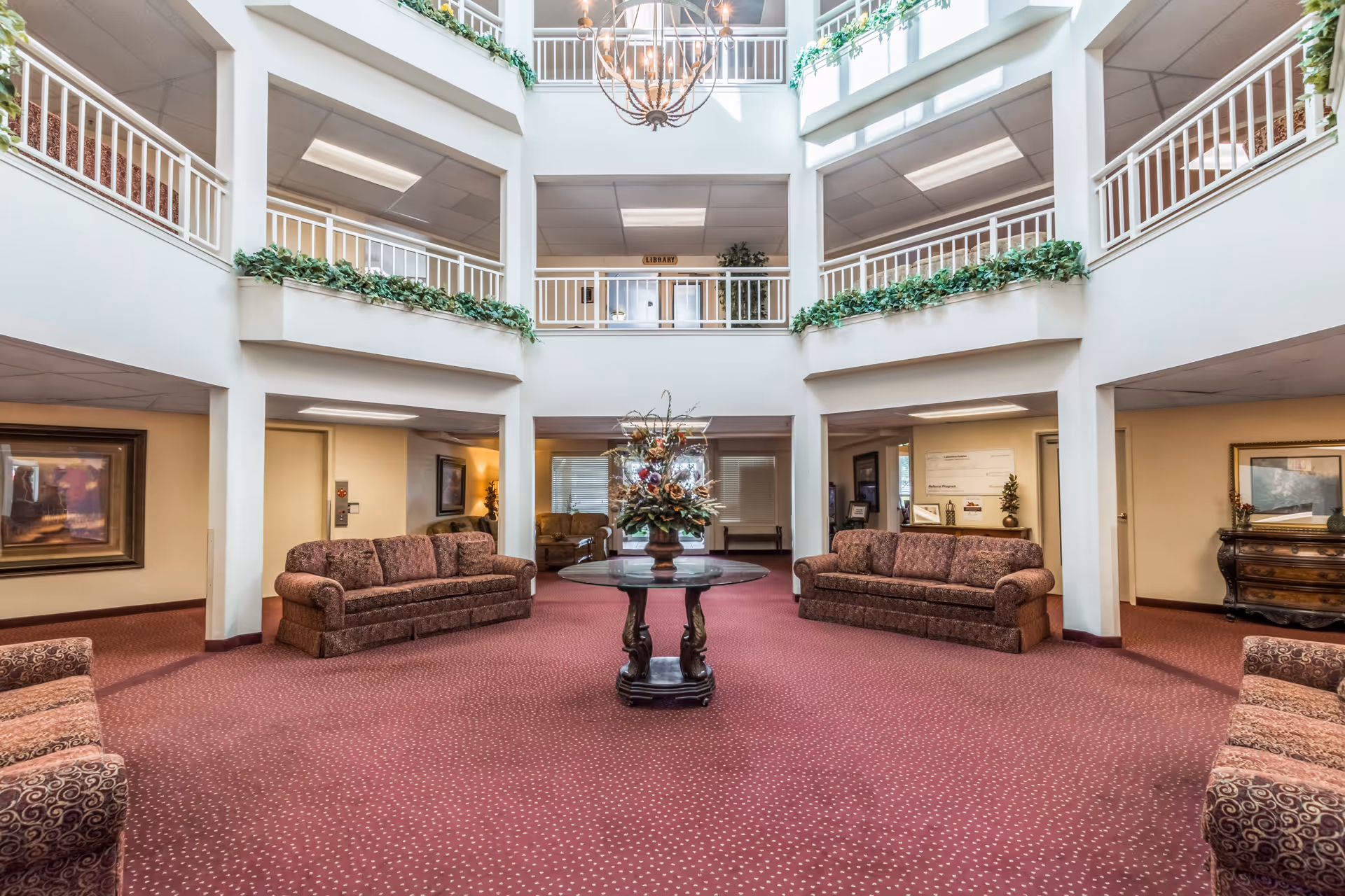 Spacious and well-lit common area in Asher Point Independent Living of Waco featuring a central round table with a floral arrangement, surrounded by multiple upholstered sofas. The room has a high ceiling with a chandelier, white railings on the upper level, and green plants decorating the balcony edges. The carpet is red with a subtle pattern, and there are framed artworks and furniture along the walls.