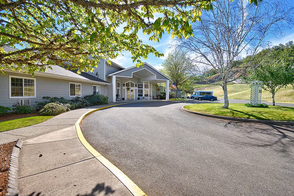 Exterior view of Bridgewood Rivers Assisted Living facility entrance with a curved driveway, sidewalk, green lawn, trees, and a clear blue sky.